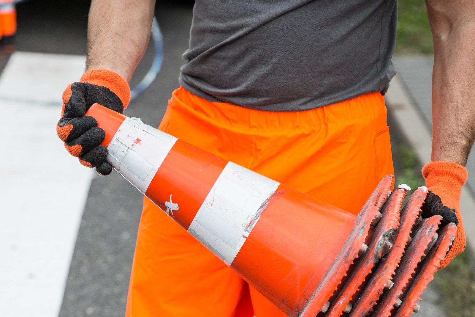 Gloved hands of a field worker in high-visibility gear holding a taped traffic cone and cone bases above painted road markings