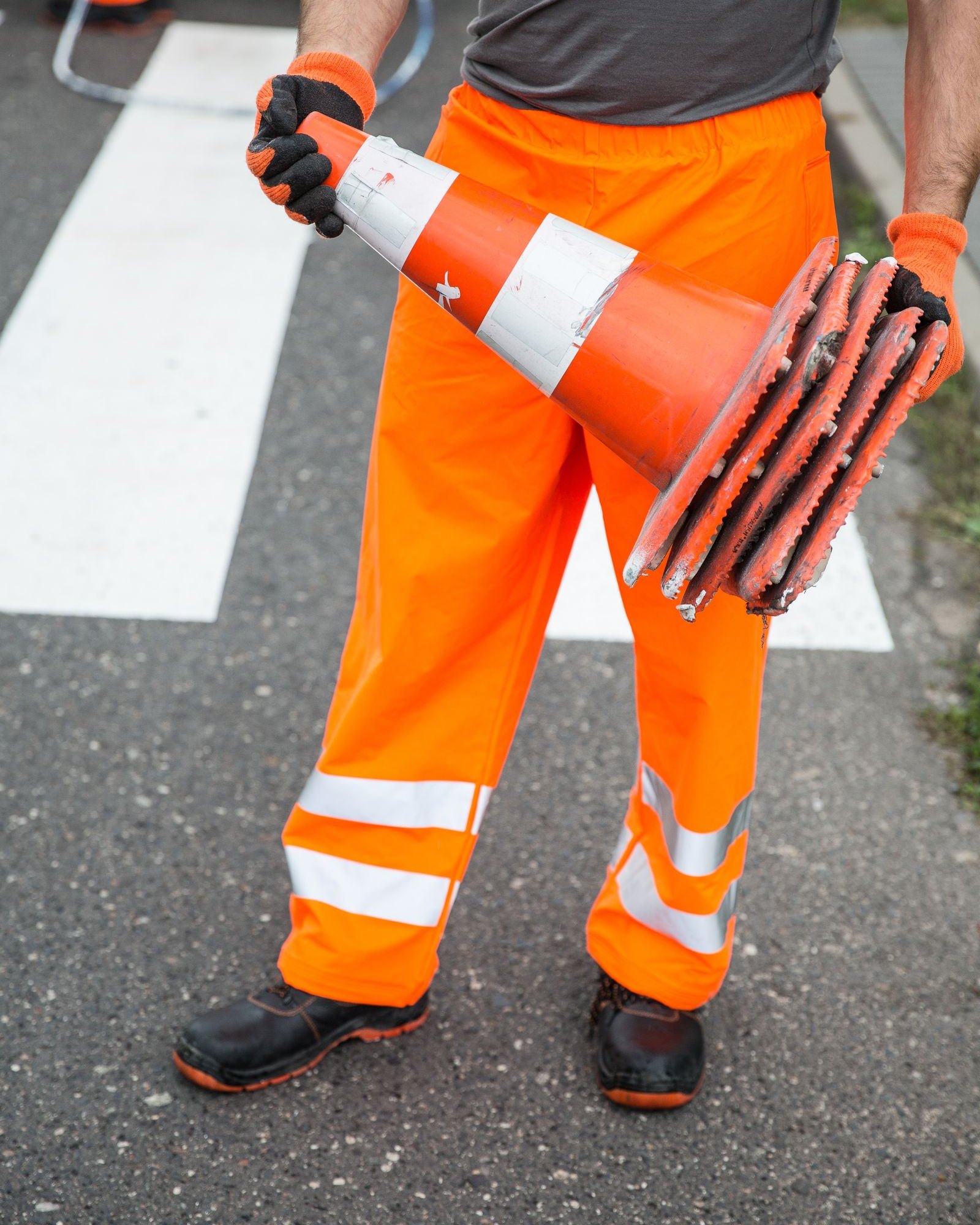 Traffic-control worker in high-visibility orange trousers and gloves carrying a stack of cone bases across a marked crossing