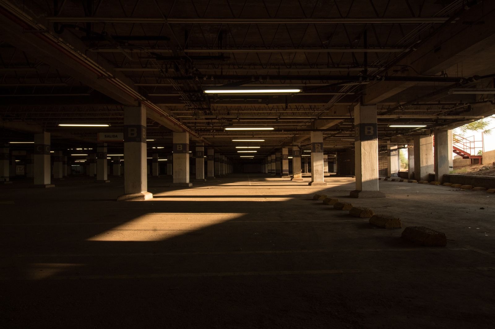 Interior of a parking structure with lettered columns, wheel stops and sunlight raking across empty bays